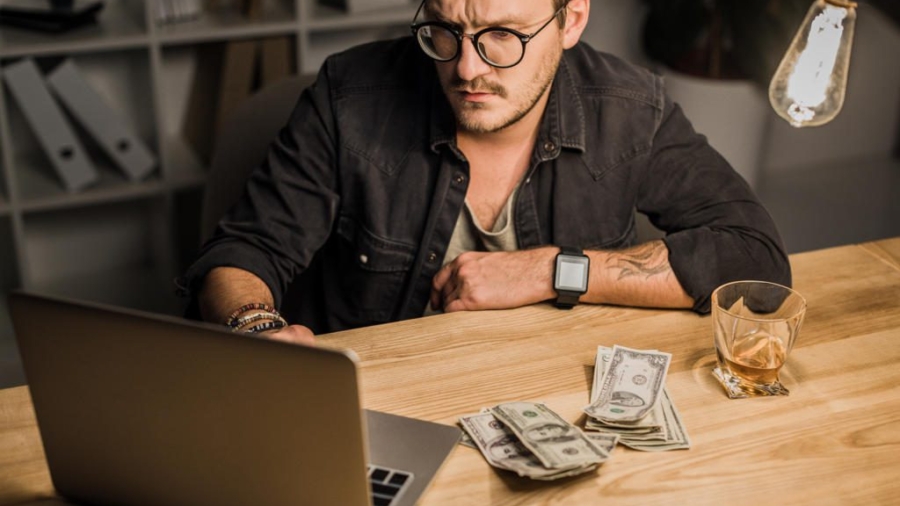 young man with whiskey and pile of cash using laptop