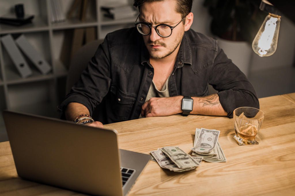young man with whiskey and pile of cash using laptop